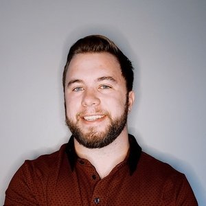 A man with short brown hair and a beard, wearing a burgundy button-up shirt, stands against a plain light gray background, smiling at the camera.