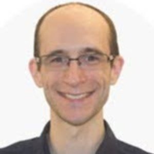 A person with short brown hair and glasses is smiling at the camera against a plain, light-colored background.