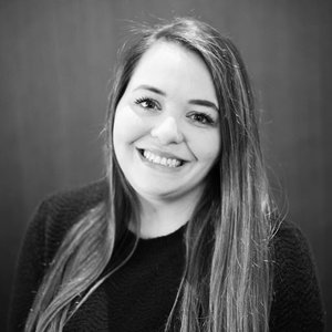 A woman with long straight hair, wearing a dark sweater, smiles at the camera against a plain, blurred background.