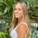 A woman with long blonde hair wearing a white sleeveless top smiles while standing in front of green leafy plants.