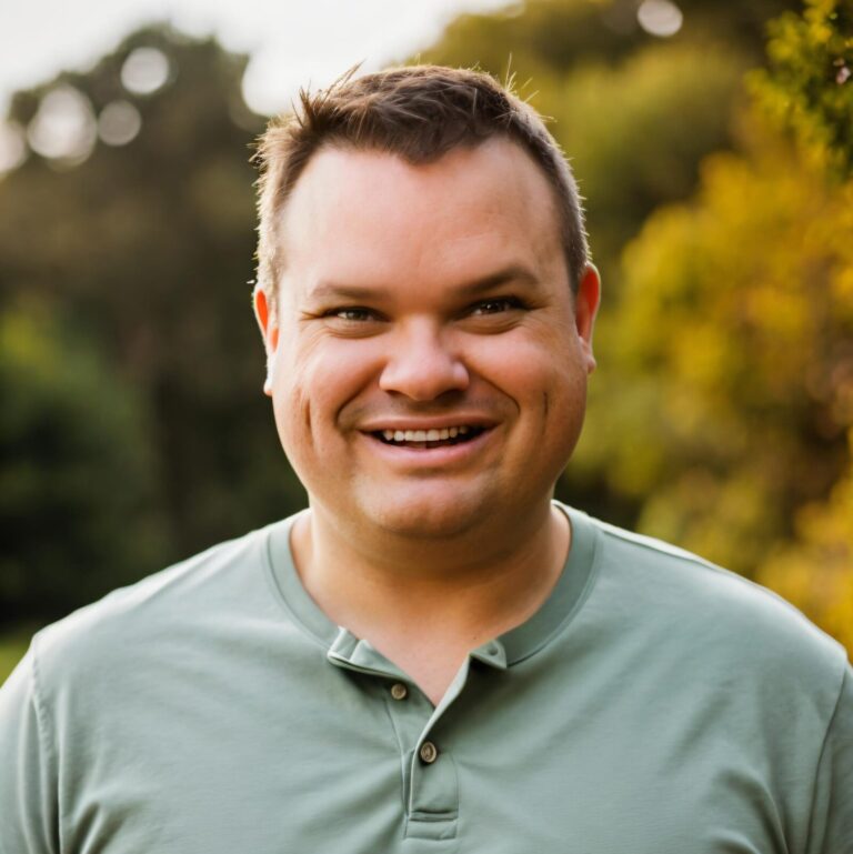 A man with short brown hair and a light green shirt smiles outdoors with trees and greenery in the background.