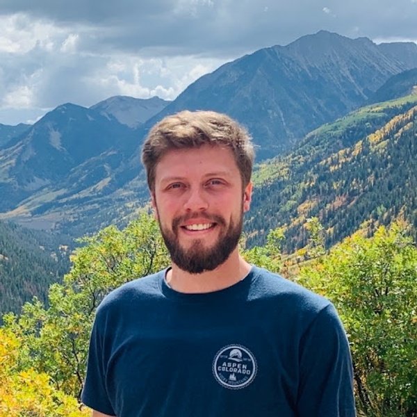 Bearded man in a navy t-shirt stands outdoors with green hills and tall mountains in the background under a partly cloudy sky.