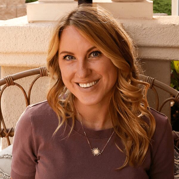 A woman with long wavy blonde hair, wearing a mauve top and a sunburst pendant necklace, sits and smiles at the camera outdoors.
