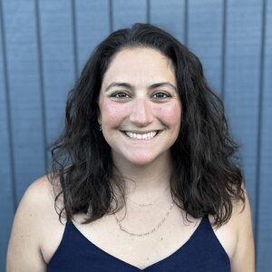 A woman with wavy dark hair and a blue tank top smiles at the camera, standing in front of a blue paneled wall.