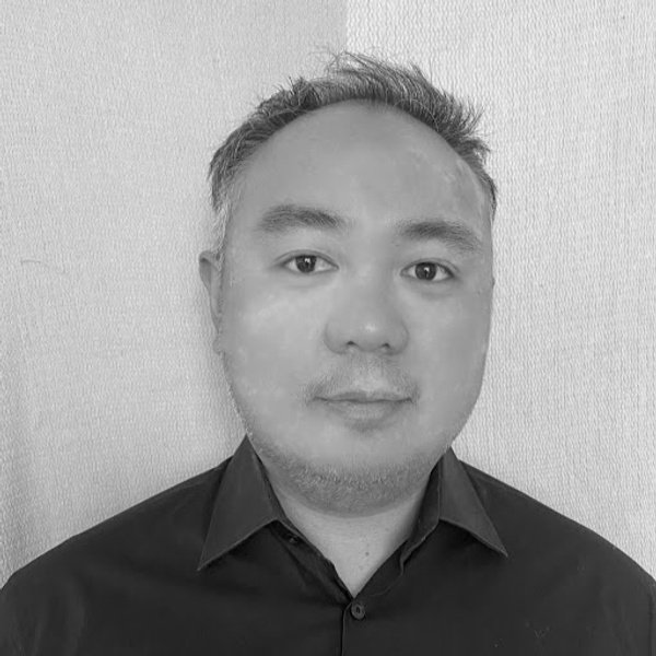 A man with short hair wearing a dark collared shirt stands in front of a light-colored textured background, facing the camera with a neutral expression.