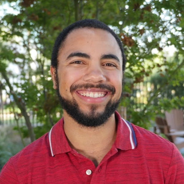 A man with short hair and a beard smiles at the camera while wearing a red collared shirt, standing outdoors in front of green trees.