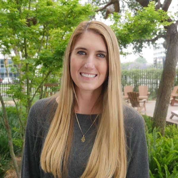 Woman with long blonde hair wearing a gray top stands outdoors in front of green trees and wooden chairs, smiling at the camera.