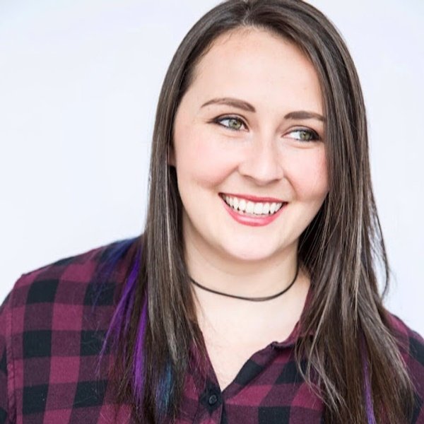 A woman with long brown hair, wearing a dark plaid shirt and a black choker, smiles while looking slightly to the side against a plain light background.