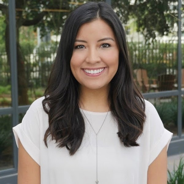 A woman with long dark hair wearing a white blouse stands outside, smiling at the camera. There are trees and a building with large windows in the background.