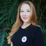 A woman with long red hair, wearing a black shirt with a white circular logo, stands in front of large green leafy plants.