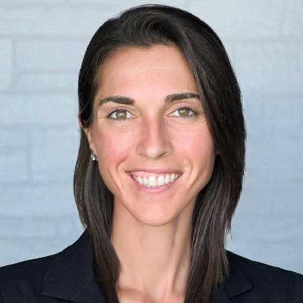 A woman with straight brown hair wearing a black blazer smiles at the camera against a light gray brick wall background.