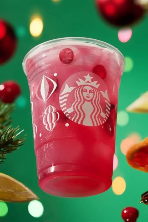 A clear Starbucks cup filled with a pink-red iced drink and ice cubes, against a festive green background with blurred ornaments and holiday decorations.