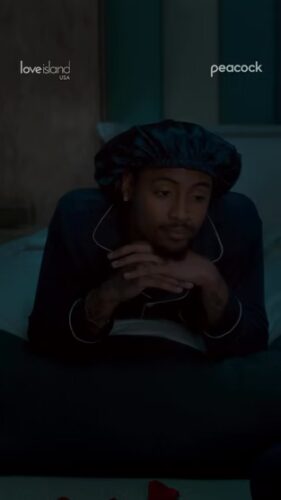 A person in dark pajamas and a satin bonnet lies on a bed with hands clasped. "Love Island USA" and "Peacock" logos appear in the top corners.