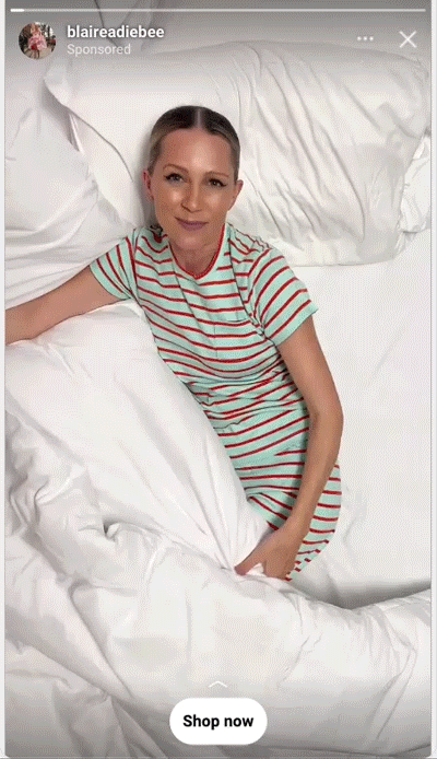 A woman wearing a striped t-shirt dress lies on a bed with white bedding, looking up at the camera. A “Shop now” button appears at the bottom of the image.
