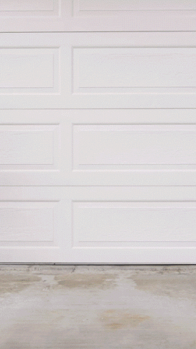 A white garage door opens to reveal a black car with bright headlights parked inside on a concrete floor.