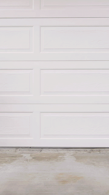A white garage door opens to reveal a black car with bright headlights parked inside on a concrete floor.