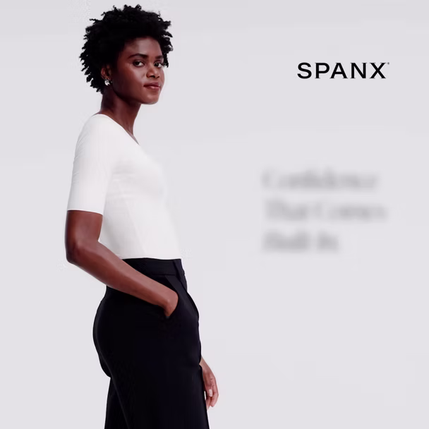 A woman wearing a white top and black pants stands with hands in pockets against a plain background. The Spanx logo appears in the upper right corner.