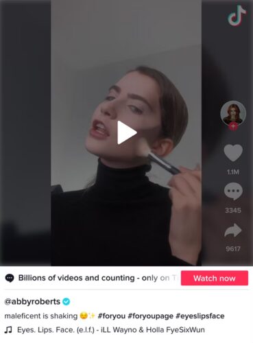 A person with slicked-back hair applies makeup to their cheek with a brush, wearing a black turtleneck and posing in front of a plain background.