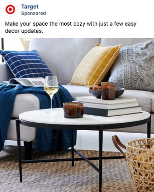 A modern living room features a white coffee table with candles, stacked books, and a glass of wine. A sofa with blue and yellow pillows and a knit blanket is in the background.
