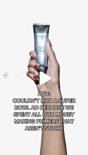 A hand holds a silver tube of Smashbox Photo Finish Primer against a plain background, with overlaid text humorously referencing Super Bowl ad spending.
