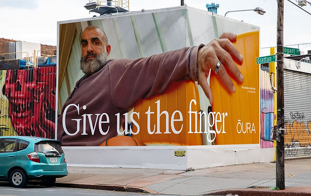 A mural on a building shows a bearded man extending his hand with a ring, next to large text that reads "Give us the finger" and the brand name "ŌURA.