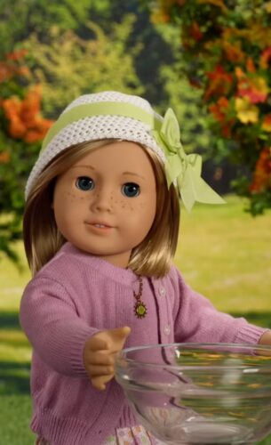 A doll with light brown hair, blue eyes, a white hat with a green bow, and a pink cardigan holds a clear glass bowl outdoors with trees in the background.