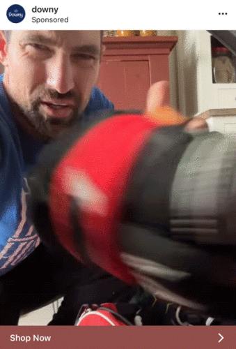 A man in a blue shirt holds a red and black athletic shoe close to the camera, partially obscuring his face.