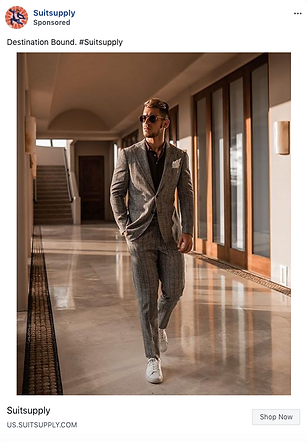 A man in a grey plaid suit and white sneakers walks confidently down a sunlit corridor with large windows.