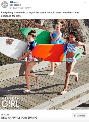 Three girls in swimsuits walk on a wooden boardwalk carrying surfboards, smiling and heading toward the beach.