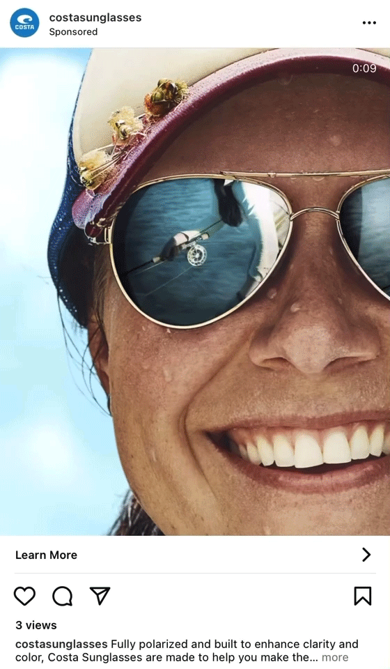 A close-up of a smiling person wearing reflective sunglasses and a hat with flowers, with a blue sky and water visible in the reflection of the lenses.