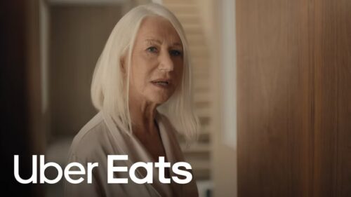 An older woman with straight white hair stands indoors, looking toward the camera. The Uber Eats logo is displayed in the bottom left corner.