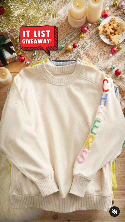 A cream-colored sweatshirt with colorful letters spelling "CHEERS" on the sleeve is displayed on a festive surface with candles, cookies, ornaments, and a "IT LIST GIVEAWAY!" sign.