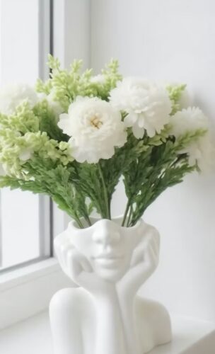 A white ceramic vase shaped like a face holds white flowers and green foliage, placed on a windowsill.