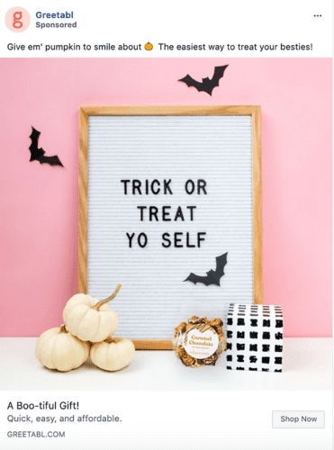A letter board reads "Trick or Treat Yo Self" against a pink wall with bat cutouts, white pumpkins, a small gift box, and caramel chocolates arranged in front.