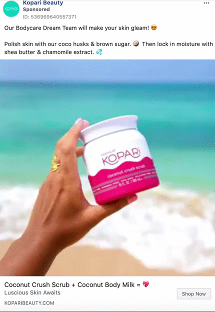 A hand with gold rings holds a jar of Kopari Coconut Crush Scrub by the beach, with turquoise water and blue sky in the background.