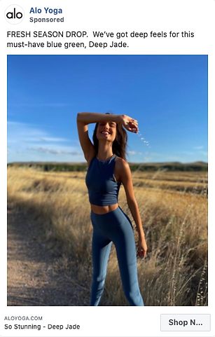 A woman stands in a field wearing a matching blue-green athletic outfit, smiling with one arm raised, under a clear blue sky.