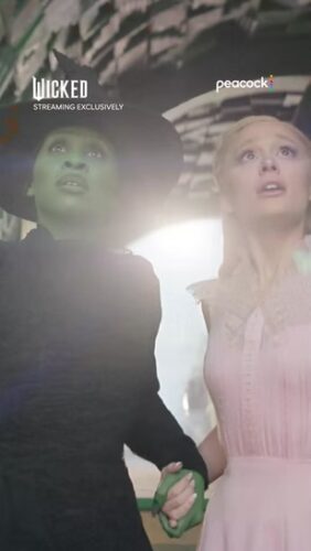 Two women, one with green skin in black attire and one in pale pink, stand close together holding hands and looking upward with concerned expressions. "Wicked" and "Peacock" logos are visible.