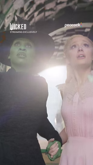 Two women, one with green skin in black attire and one in pale pink, stand close together holding hands and looking upward with concerned expressions. "Wicked" and "Peacock" logos are visible.