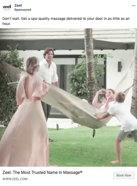 Three people hold a large blanket outdoors while a young boy jumps onto it, smiling. Modern house and greenery are visible in the background.