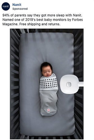 A baby swaddled in a grey blanket lies in a crib, with a white baby monitor attached to the crib rail above.