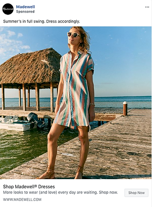 A woman wearing a striped summer dress and sunglasses stands on a wooden dock by the water with a thatched-roof hut in the background.