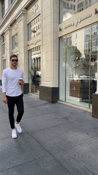 A man wearing sunglasses, a white shirt, black pants, and white sneakers walks on a city sidewalk holding a coffee cup.