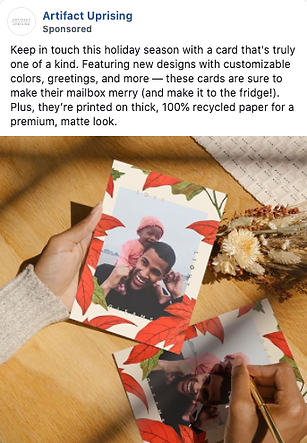 A person writes on a greeting card featuring a photo of a man and child, surrounded by autumn leaves, on a wooden table under sunlight.