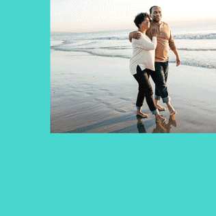 Two people walk barefoot along the shoreline, arm in arm, with waves in the background on a sandy beach.