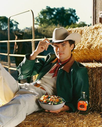A man in cowboy attire sits on hay bales, holding a bowl of salad and tipping his hat, with a bottle labeled "Nic's Maple Syrup" beside him. A horse and fence are in the background.