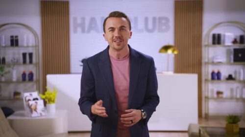 A man in a navy blazer and pink shirt stands in a modern, well-lit room with decorative shelves and "HAIR CLUB" signage in the background.