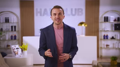 A man in a dark blazer and pink shirt stands indoors in front of a reception desk with "HAIR CLUB" on the wall behind him. Shelves with products and plants are visible.