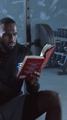 A man sits in a gym in front of a loaded barbell, reading a book while wearing athletic clothing.