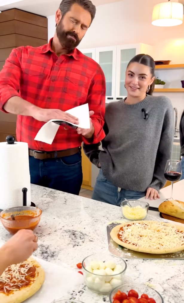 A man wipes his phone with a paper towel while a woman stands next to him, both in a kitchen with pizzas and pizza ingredients on the counter.