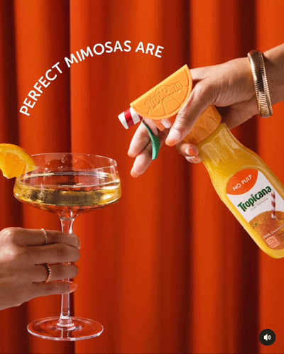 A hand sprays Tropicana orange juice from a spray bottle into a cocktail glass with an orange slice garnish, against a red curtain background. Text reads, "PERFECT MIMOSAS ARE.